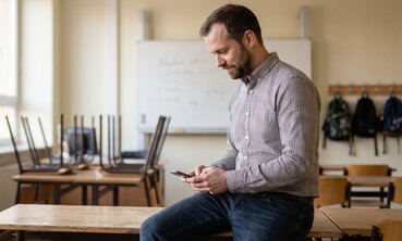 Lehrer schreibt nach Unterrichtsschluss im leeren Klassenzimmer eine Nachricht auf dem Smartphone